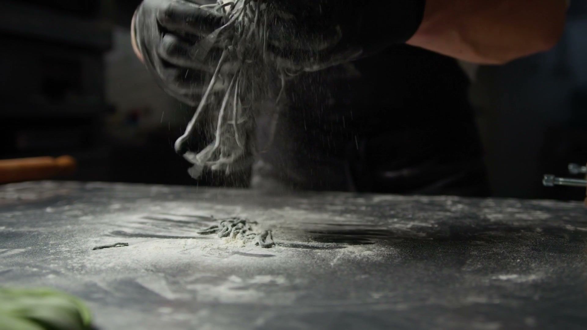 Gloved hands shaking flour off fresh dark pasta over a floured steel counter
